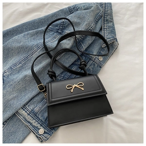 Detail of a black leather crossbody bag featuring a golden metal bow buckle on a denim background