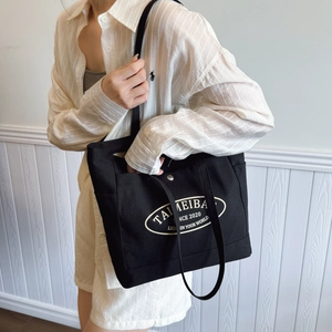 Black canvas tote bag with white logo and snap closure worn on shoulder by a model