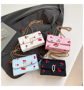 Flat lay of four cherry-print crossbody bags in white, pink, blue, and black with gold chains