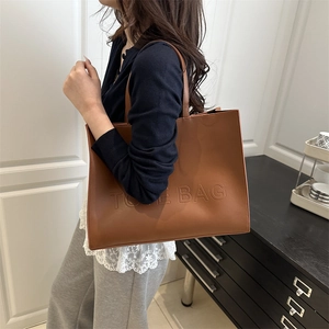 Brown faux leather tote bag with embossed letters worn by a model in a modern room