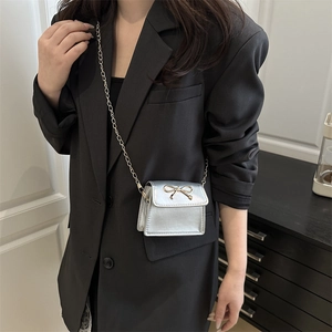 Silver mini crossbody bag with gold bow detail and chain strap worn on black blazer