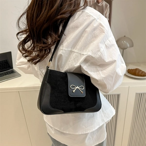 Minimalist black suede shoulder bag with a gold bow buckle worn by a model in a modern room