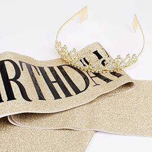 Close-up of golden birthday sash with black text and sparkling rhinestone crown on white background