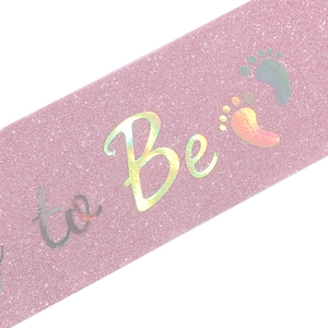 Detail of pink glitter sash with holographic to Be text and baby footprints for baby shower
