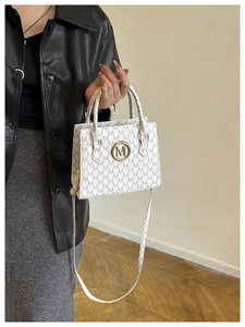 Detail der weißen Tasche mit M-Muster, goldenem Logo und verstellbarem Schulterriemen
