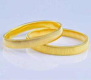 Detail view of two gold metal spring armbands with vertical textures on a gradient base