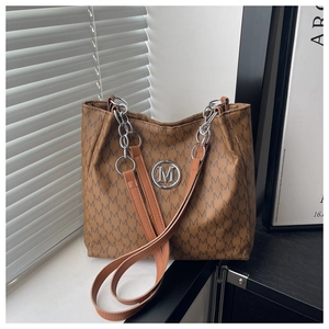 Detail of a brown tote bag featuring M-letter pattern and a silver round logo