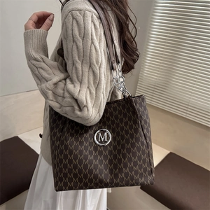 Brown monogram tote bag with metallic logo and chain strap worn by a woman in cozy sweater