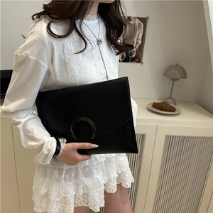 Minimalist black envelope clutch bag with moon-shaped clasp held by model in white dress