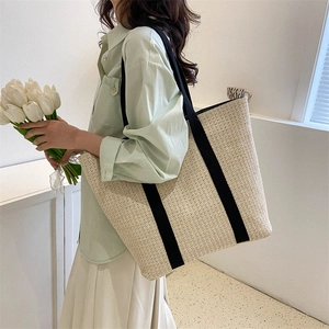 Large straw tote bag with black canvas straps shown on model in a cozy indoor setting