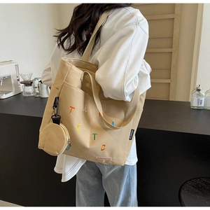 Side view of beige canvas tote bag featuring letter embroidery and small coin pouch charm