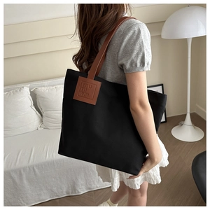 Close-up of black canvas tote bag with brown leather handles worn by a model in a room