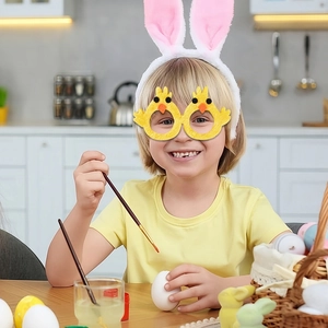 Kid wearing yellow chick Easter glasses frame while decorating colorful festive eggs