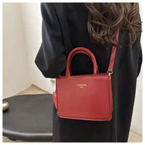 Détail du sac carré en cuir rouge avec lettrage doré porté sur un blazer noir élégant