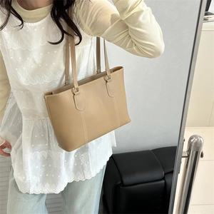 Minimalist khaki leather tote bag with contrast stitching, worn by a model in a warm room