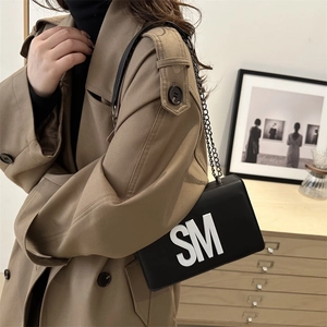 Petit sac bandoulière en cuir noir avec lettres SM porté sur un trench-coat kaki élégant