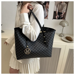 Detail of black tote bag with M-pattern print and golden logo charm on a shoulder strap