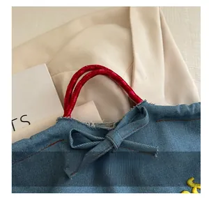 Detail of blue denim tote bag featuring red braided handles and a decorative denim bow