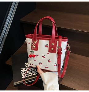 Close-up of white bucket bag with cherry print, red handle, and charm on wooden stairs