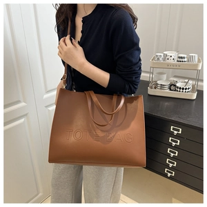 Brown leather tote bag featuring embossed TOTE BAG text and adjustable strap on a model
