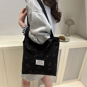 Black corduroy tote bag with cherry print and wide straps on female model in home setting