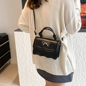 Chic black crossbody bag with scalloped flap and gold bow accent on a stylish model