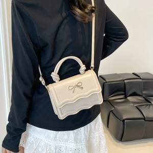 Beige crossbody mini bag with wavy flap design and golden bow detail on a model