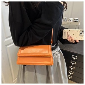 Bright orange rectangular crossbody bag shown worn by a model in a minimalist home setting