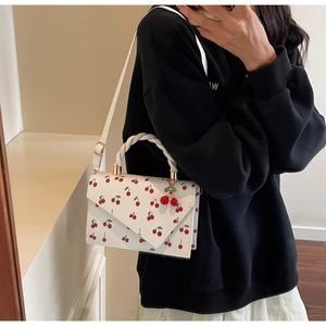 Detail of white square crossbody bag with red cherry print and twisted top handle worn by model