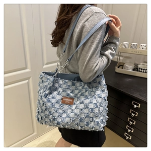Detail of blue plaid tote bag with raw edges and a brown leather logo patch on shoulder