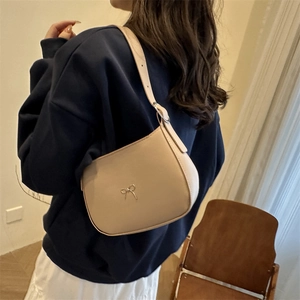 Minimalist beige crossbody bag with gold bow clasp shown on model in warm indoor setting