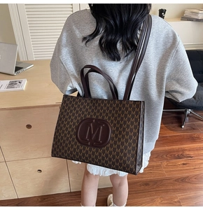 Detail of large brown tote bag with all-over M pattern and shoulder strap in office setting