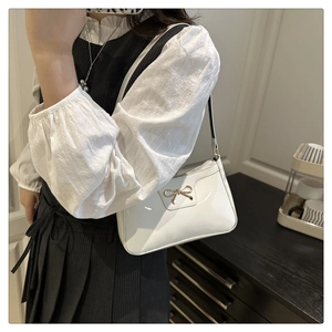 Close-up of a white patent leather baguette bag with a gold metal bow on a shoulder strap