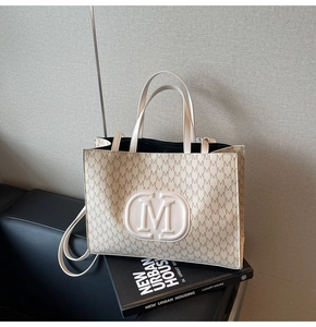 Detail of beige tote bag featuring letter M pattern and embossed logo on leather surface