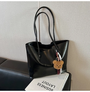 Detail of black glossy faux leather tote bag with a brown plush bear charm on a sofa