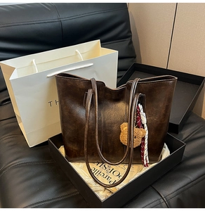 Detail of dark brown leather tote bag featuring a teddy bear charm and woven strap accents