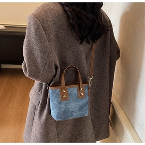Blue suede bucket bag with brown leather handles shown worn by a model on a dark coat