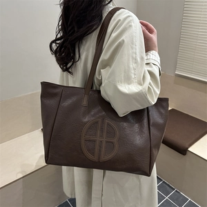 Large brown leather tote bag with round letter stitching shown in a minimalist studio setting