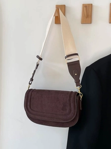 Brown suede saddle bag with a wide beige strap hanging on a wooden hook in a minimalist setting