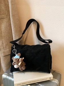 Quilted black shoulder bag with wide strap and teddy bear charm on a silver suitcase