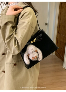 Detail of matte black bucket bag with gold lock and plush rabbit charm on a trench coat