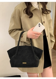 Detail of a black suede tote bag with a golden metal bar held by a model in a beige coat