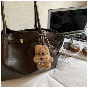 Close-up of a light brown plush dog charm with silver bone tag on a leather tote bag