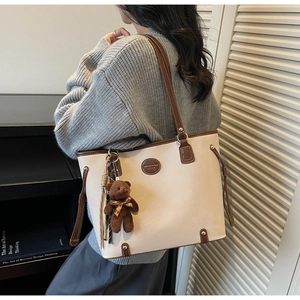 Detail of a beige tote bag featuring a brown teddy bear charm and leather tassel accents