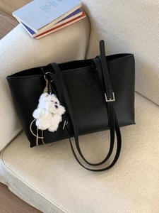 Large black leather tote bag with adjustable strap and cute plush puppy charm on a sofa