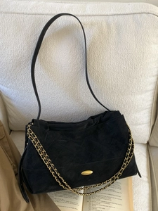 Black suede tote bag with chain strap and gold logo displayed on a textured sofa