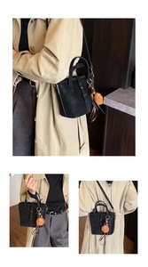 Détail d'un sac seau en cuir noir avec un porte-clé en peluche marron porté sur un trench