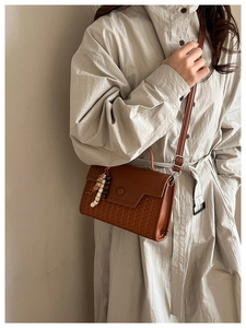Close-up of brown leather crossbody bag with woven texture and bead charm on a beige coat