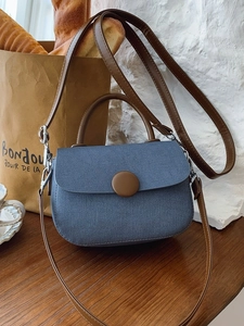 Chic blue denim crossbody bag with brown leather handles on a French-style wooden table