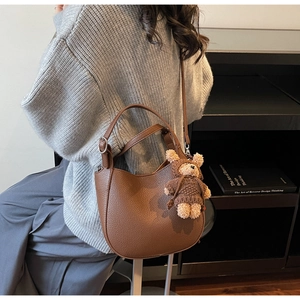 Braune Handtasche mit Litschi-Struktur und Teddybär-Anhänger auf grauem Strickpullover